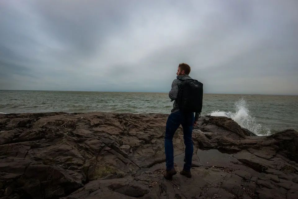 Man standing on rocky shore in Duluth, Minnesota, facing the lake under overcast sky.