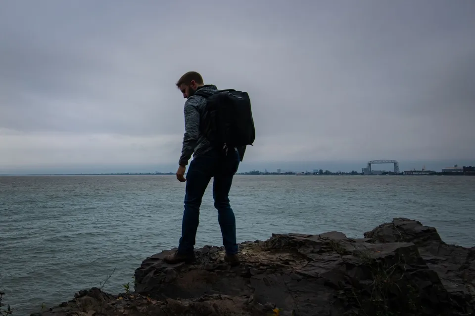 A man with a backpack stands on rocky shore, gazing over Duluth's waterfront.