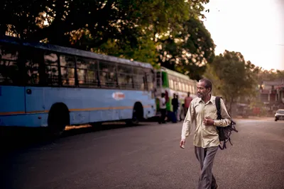 An adult man walks beside a bus on a tree-lined urban street during sunset, carrying a backpack.