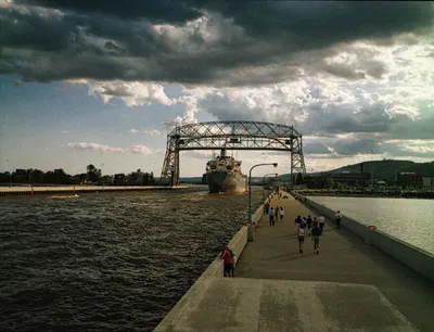 A scenic view of Duluth's Aerial Lift Bridge with ship passing and people strolling on a cloudy day.