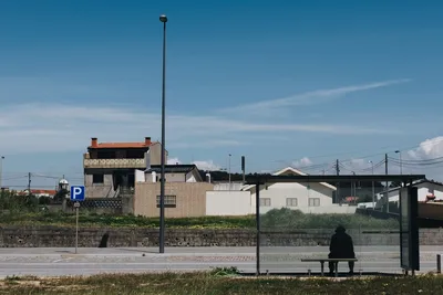 A solitary figure sitting at a bus stop in a rural area, under a bright blue sky.
