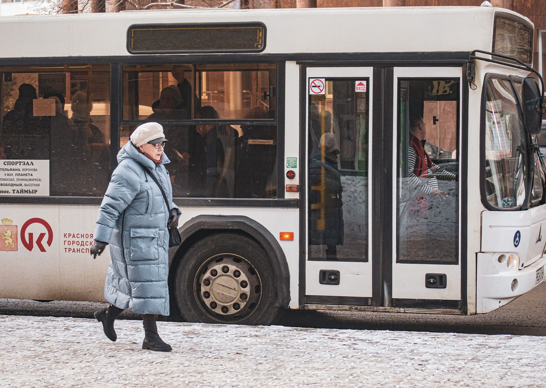 A woman in a winter coat walks past a bus on a snowy city street, capturing urban life.