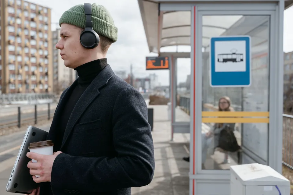 A man in a black coat and green beanie waits at a bus stop, holding coffee and a laptop.